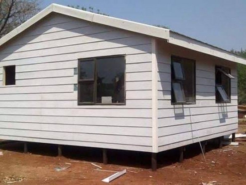 Small white Nutec house with a simple rectangular structure, featuring horizontal siding, a pitched roof, and dark-framed windows. The house is elevated on short stilts, with open windows for ventilation, set on a bare outdoor ground area.