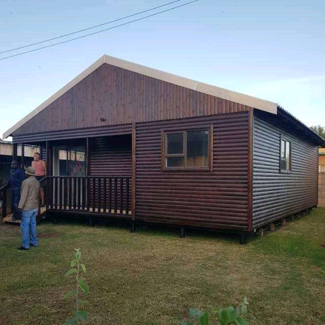 Mutsamvi Wendy houses A large wooden Wendy house elevated on stilts with a front porch, railing, and steps leading up to the entrance. The house features multiple windows and a pitched roof, set in a dry, open landscape under a clear sky, providing a rustic and spacious design ideal for extended outdoor living.
